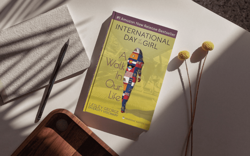 International Day Of The Girl - A walk in our Life: Pandemic Edition A copy of the book on a table with minimal neutral decorations. There are some shadows indicating a nearby window, with sunlight shining on the book in the center of the image.